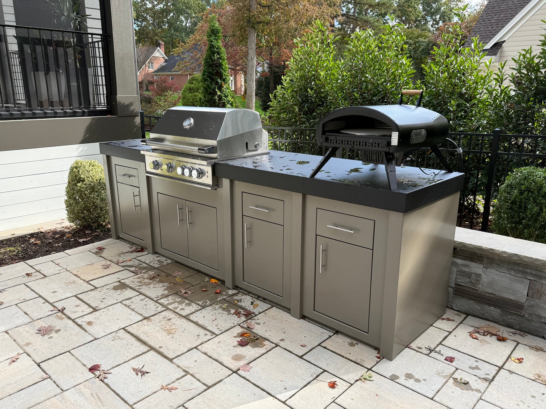 A taupe Stono engineered outdoor kitchen with a built-in stainless grill and a black kamado-style grill on the counter, set on a stone paver patio beside a house.