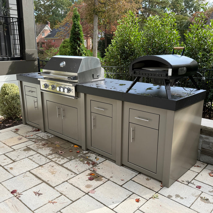A taupe Stono engineered outdoor kitchen with a built-in stainless grill and a black kamado-style grill on the counter, set on a stone paver patio beside a house.