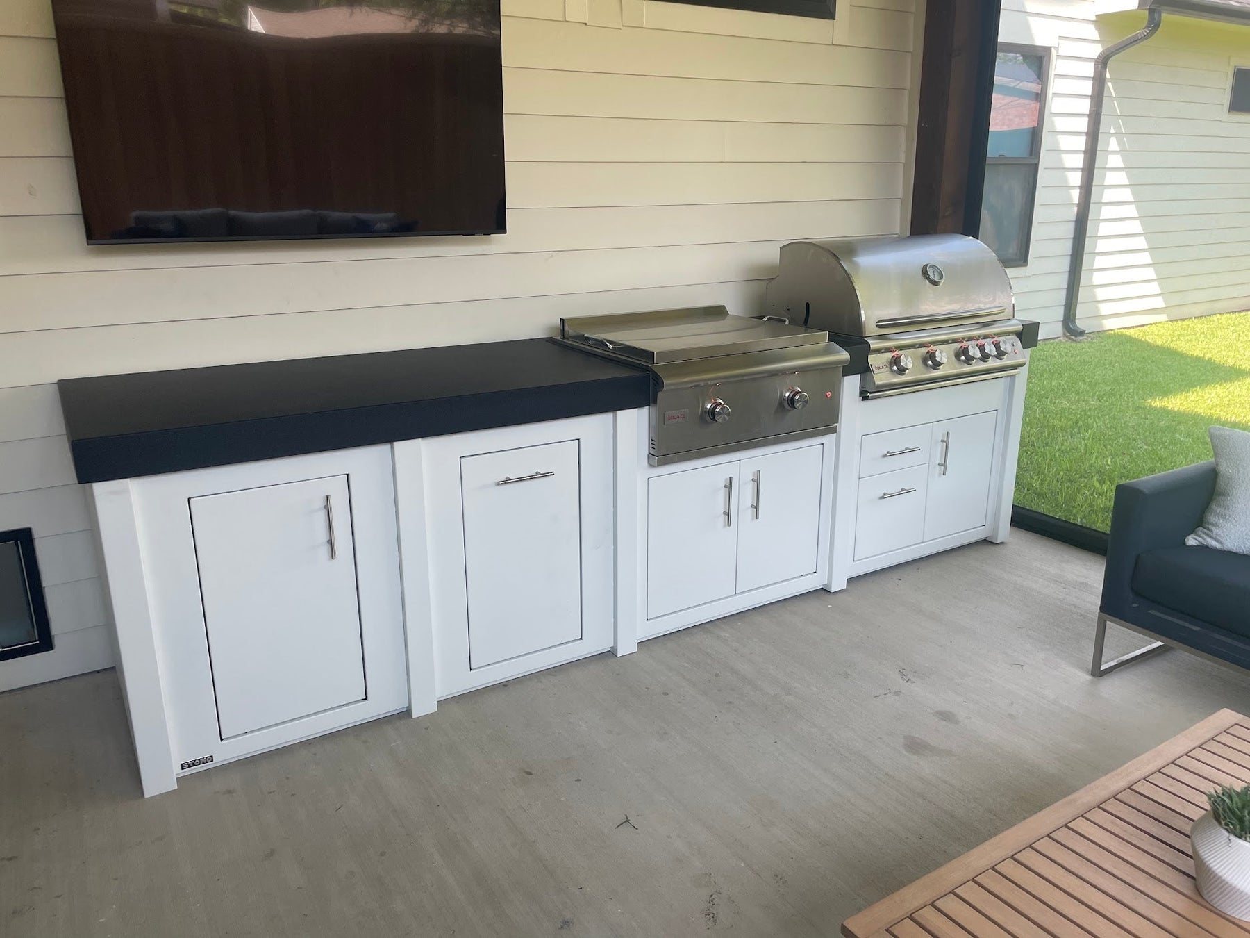 A long white Stono engineered outdoor kitchen with a dark countertop and two built-in stainless grills, set under a covered patio with a TV mounted on the wall above.