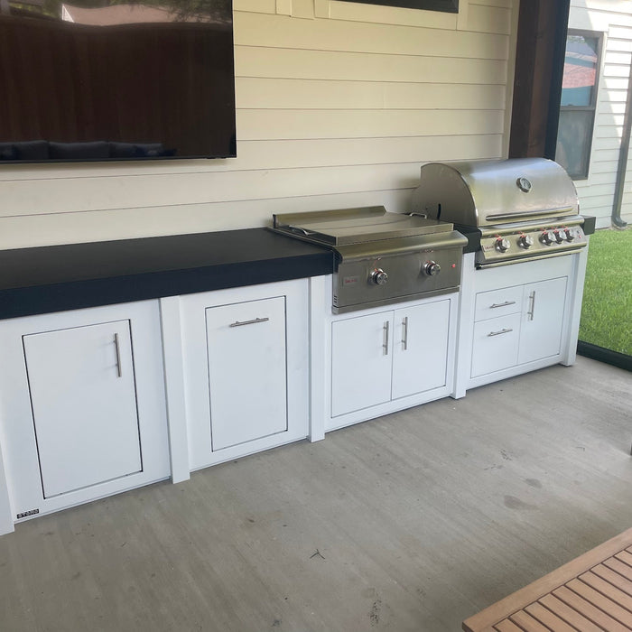 A long white Stono engineered outdoor kitchen with a dark countertop and two built-in stainless grills, set under a covered patio with a TV mounted on the wall above.