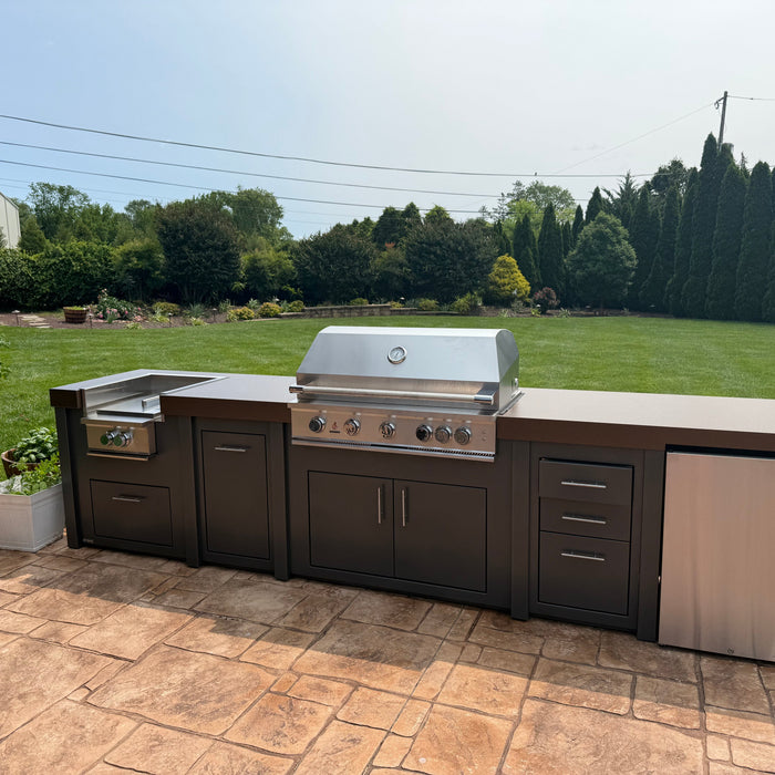 A Stono engineered outdoor kitchen with a built-in stainless grill and storage cabinets on an open patio, exposed to the weather.